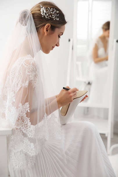 Young bride writing her single friends names on shoe indoors. Wedding superstition - Foto, Bild