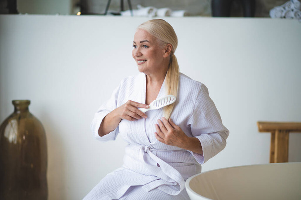 Usmívající se potěšená blondýnka oblečená v froté županu sedí v koupelně a kartáčuje si vlasy - Fotografie, Obrázek