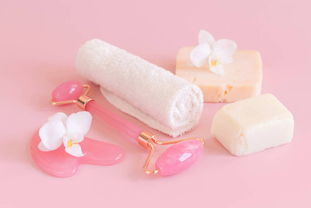Facial roller, gua sha massager, towel and handmade soap bars near white orchid flower on pink close up. Anti-age massage and natural lifting therapy at home. Facial skin care at home. Beauty concept - Photo, Image