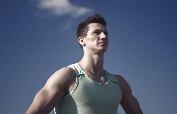 Athlete in green tshirt on sunny day. Fitness model with muscular hands, biceps, triceps, strong muscles. Workout and training. Sport and healthy lifestyle concept. Man bodybuilder posing on blue sky. - Photo, Image