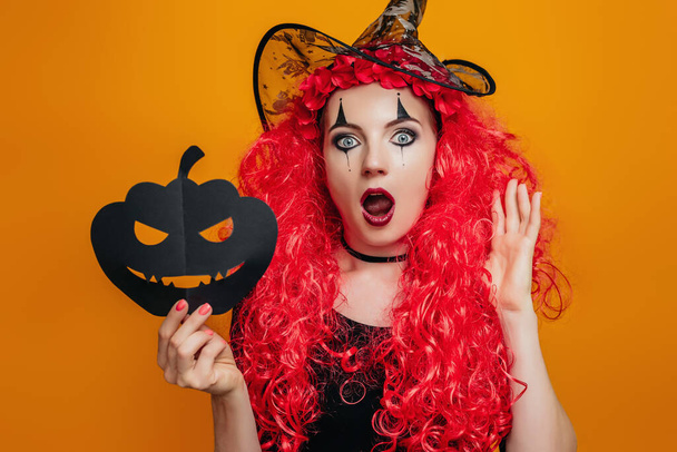 Chica payaso sorprendido en traje de Halloween sostiene una calabaza de papel y grita. Modelo femenino conmocionado con calabaza de papel negro en manos aisladas sobre fondo naranja - Foto, Imagen