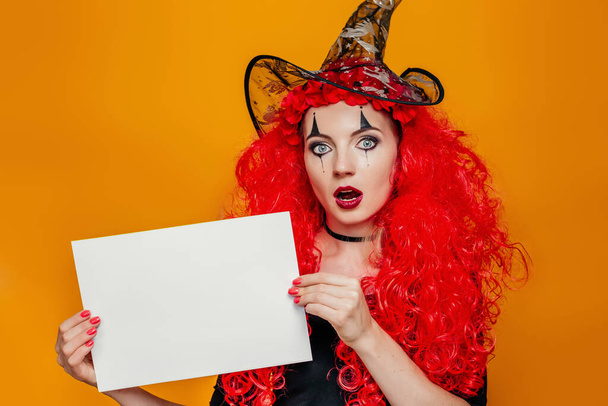 Vrouw in Halloween kostuum met een wit vel papier. Geschokt vrouwelijke clown in een rode pruik met make-up toont een A4 poster. Kopieer ruimte voor tekst - Foto, afbeelding