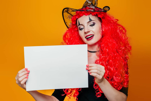 Femme en costume d'Halloween tenant une feuille blanche de papier. Un clown féminin souriant avec une perruque rouge et un maquillage montre une affiche A4. Espace de copie pour le texte - Photo, image