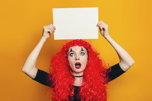 Een meisje in een Halloween kostuum houdt een vel papier boven haar hoofd. Jonge geschokte vrouw in rode krullende pruik met wit vel papier en kijken naar het geïsoleerd op gele achtergrond - Foto, afbeelding