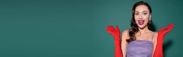 amazed young woman with red lips and gloves gesturing on green background, banner - Foto, imagen