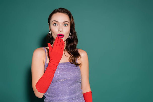 wstrząśnięta młoda kobieta w czerwonych rękawiczkach i fioletowej sukience pokrywającej otwarte usta na zielono - Zdjęcie, obraz