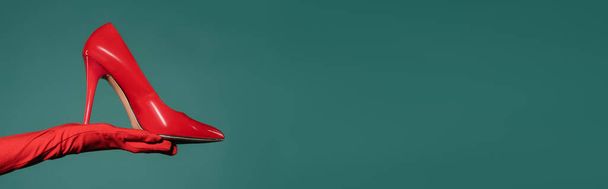 cropped view of woman in red glove holding high heeled shoe on green background, banner - Foto, imagen