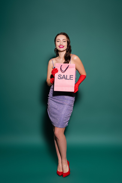 happy young woman in red gloves and purple dress holding shopping bag with sale lettering on green  - Photo, Image