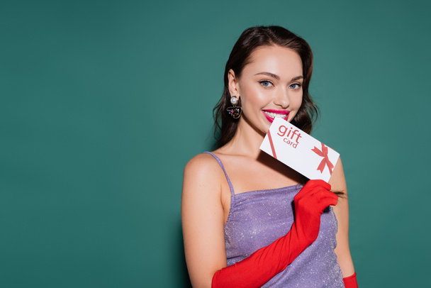 cheerful young woman in red gloves and purple dress holding gift card on green  - Foto, Bild