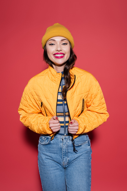 happy young woman in beanie hat adjusting puffer jacket and smiling on red - Foto, imagen