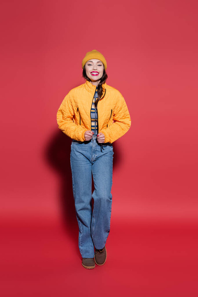 full length of happy young woman in beanie hat adjusting orange puffer jacket and smiling on red - 写真・画像