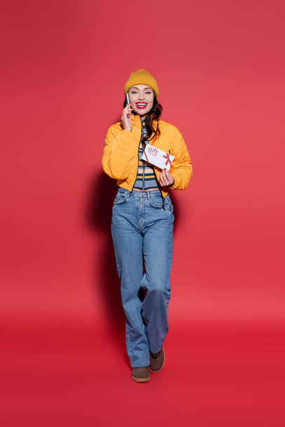 full length of cheerful woman in beanie hat and puffer jacket holding gift card while talking on smartphone on red - Photo, image