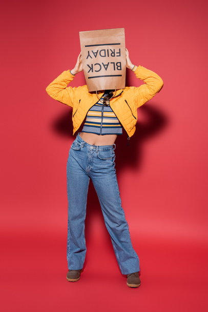 full length of woman in orange puffer jacket with black friday paper bag on head standing on red - Foto, Imagem