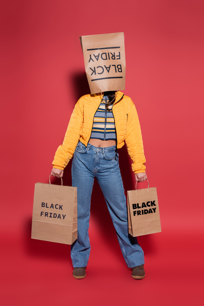 full length of woman in orange puffer jacket with black friday paper bag on head standing with shopping bags on red - Fotoğraf, Görsel