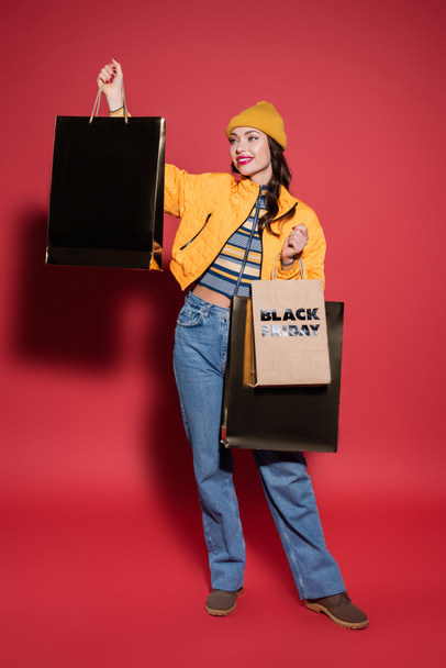 full length of cheerful woman in beanie hat and puffer jacket holding shopping bags with black friday lettering on red - Foto, Bild