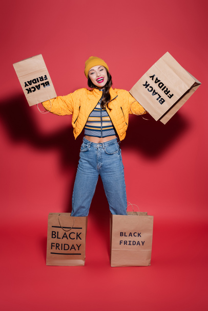 cheerful woman in beanie hat and puffer jacket with black friday shopping bags on hands standing on red - 写真・画像