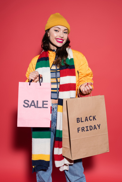 joyful woman in beanie hat and scarf holding shopping bags with black friday lettering on red  - Foto, imagen