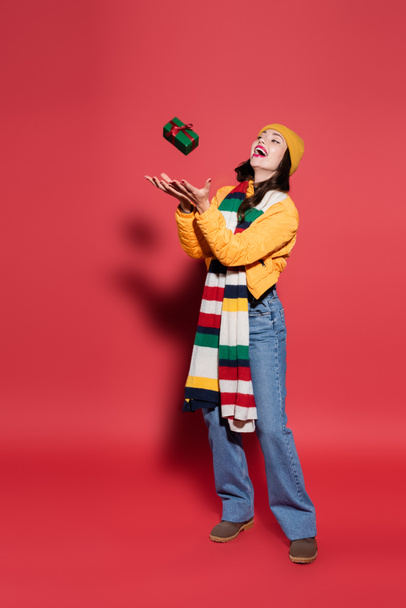 full length of amazed woman in beanie hat and scarf catching wrapped gift box on red - Foto, imagen