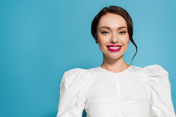 charming young woman in white blouse smiling while looking at camera isolated on blue - Foto, afbeelding