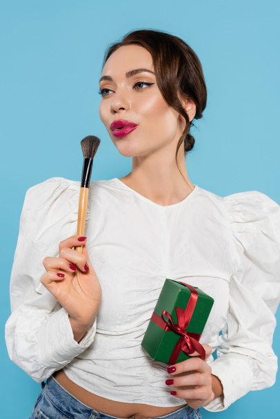 brunette woman in white blouse holding wrapped present and blowing on cosmetic brush isolated on blue - Fotoğraf, Görsel