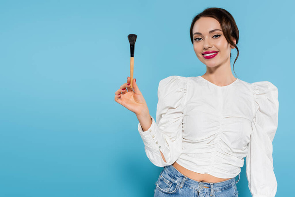 cheerful young woman in white blouse holding cosmetic brush and smiling on blue - Foto, Imagem