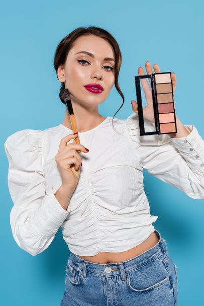 young woman in white blouse holding cosmetic brush and eye shadow palette isolated on blue - 写真・画像