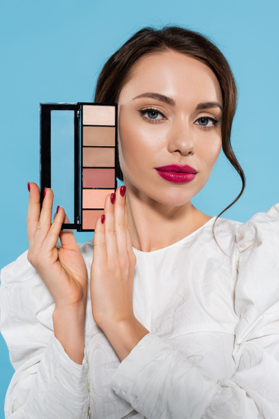 pretty young woman in white blouse holding eye shadow palette isolated on blue  - Photo, Image
