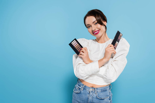 happy young woman in blouse holding eye shadow palettes on blue background - Photo, Image