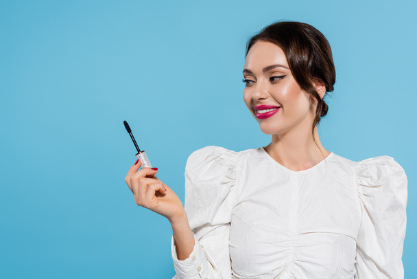 cheerful young woman in white blouse holding mascara brush isolated on blue - Foto, immagini