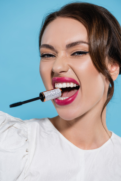 brunette young woman in white blouse holding mascara brush in teeth isolated on blue - Foto, afbeelding