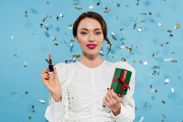 cheerful young woman in white blouse holding lipstick and gift box near falling confetti on blue - Photo, Image
