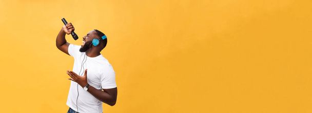 Портрет веселого позитивного шика. Красивый африканский мужчина с микрофоном и наушниками на голове, слушающий музыку, поющий песню, наслаждаясь отдыхом в выходные дни изолированы на желтом фоне. - Фото, изображение