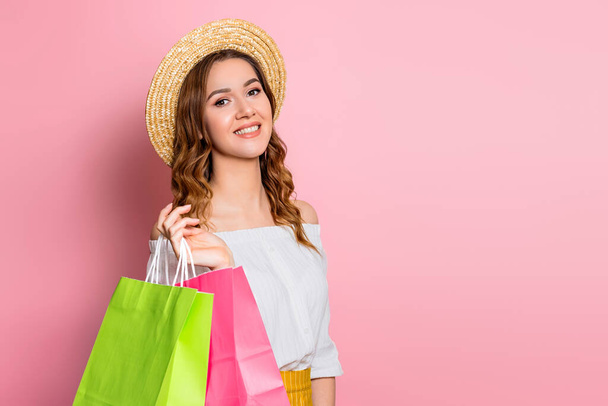 Mladá dívka s vlnitými vlasy v slaměném klobouku a retro oblečení se zelenými a růžovými nákupními taškami na růžovém pozadí, kopírovací prostor. Veselá dívka se usmívá, nakupuje a dívá se do kamery - Fotografie, Obrázek