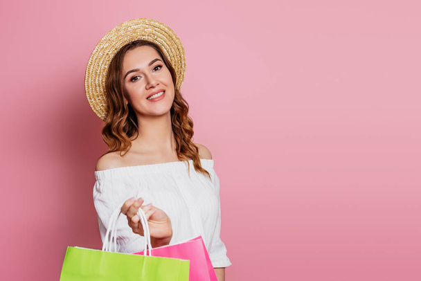 Mujer joven con el pelo ondulado en un sombrero de paja y vestido vintage con un rosa y verde bolsas de compras sobre un fondo rosa, espacio de copia. Chica sonríe y hace compras en línea, concepto de venta - Foto, imagen
