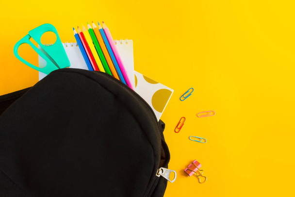 Чорний рюкзак і шкільне приладдя на жовтому тлі з копіювальним простором, плоский. Кольорові олівці, ножиці, рюкзак, паперові затискачі, папір на звичайному тлі
 - Фото, зображення