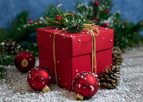 A close-up vertical photo of a red color gift box that stands on a wooden table among Christmas tree toys and coniferous branches against a wall with an abstract blue color texture. - Photo, image