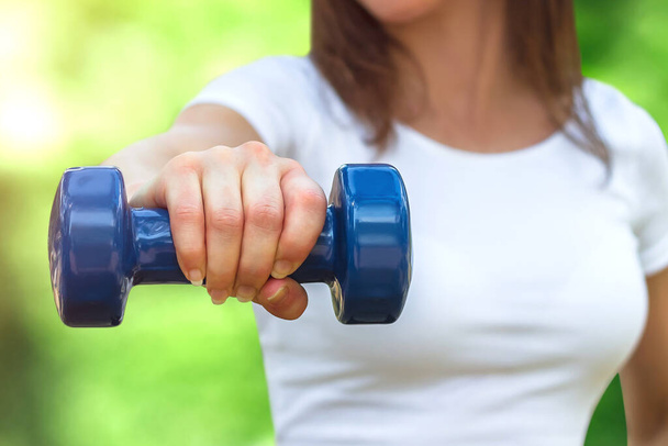 Sport training concept. Female hand is holding a blue dumbbell against the background of the park. The girl is engaged in fitness in nature - Photo, Image