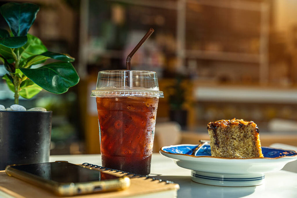 Americano iced coffee or black coffee in cup and Homemade Banana Cake, Cashew Nut Topping with smartphone on notebook on wood desk on top view In a coffee shop at the cafe,during business work concept - Foto, Imagem