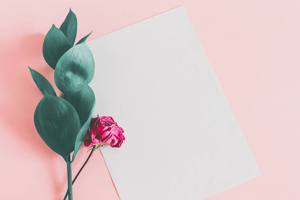 White sheet of paper on a pink background with green leaves and a dry pink rose top view mockup. Floral background for lettering with copy space. - Photo, Image