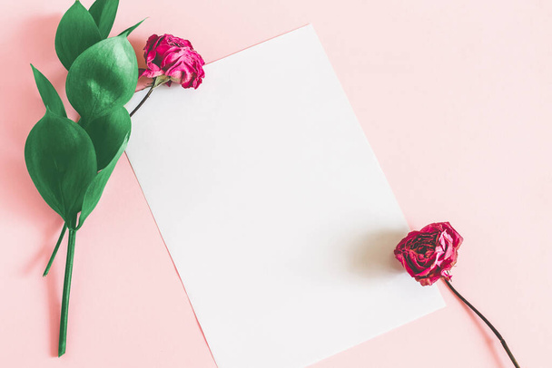 Wit vel papier op een roze achtergrond met groene bladeren en een droge roze roos bovenaanzicht mockup. Bloemen achtergrond voor belettering met kopieerruimte. - Foto, afbeelding
