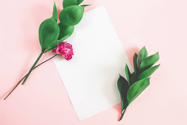 Fehér papírlap rózsaszín háttérrel, zöld levelekkel és száraz rózsaszín felülnézeti mockup. Virágos háttér betűk másolási hely. - Fotó, kép
