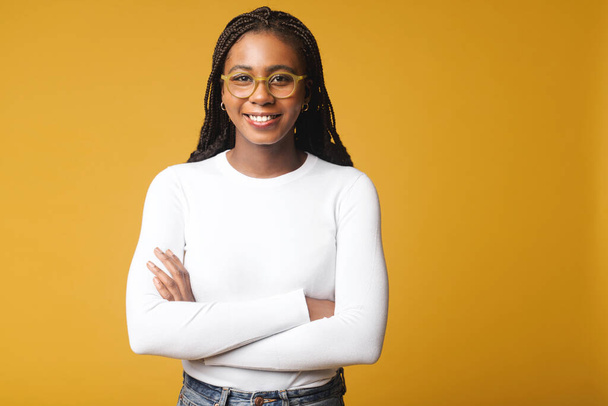 Happy young multiracial woman with folded arms stands near empty space. Cheerful female youngster wearing white t shirt with arms crossed isolated yellow colour background - Foto, imagen