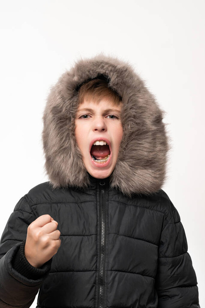 A boy in a winter jacket with a hood on a white background, showed the aggression of a child in emotions, the cry of a child from anger. - Foto, afbeelding