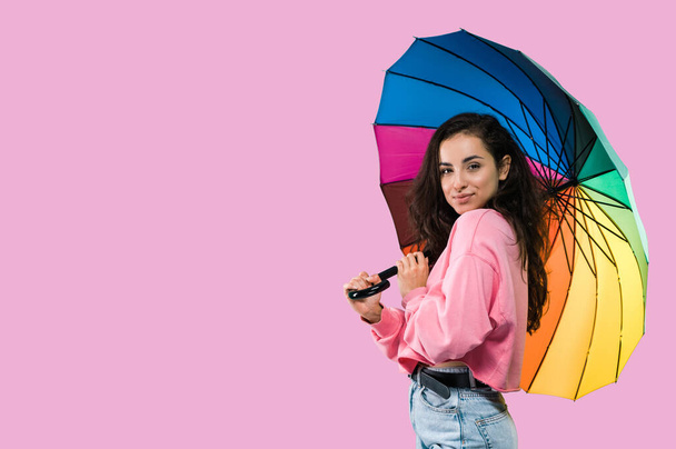 Beautiful stylish young caucasian woman with long hair, standing over isolated pink background by rainbow colored umbrella, looking at camera, smiling. Copy space. Space for presentation - Foto, imagen
