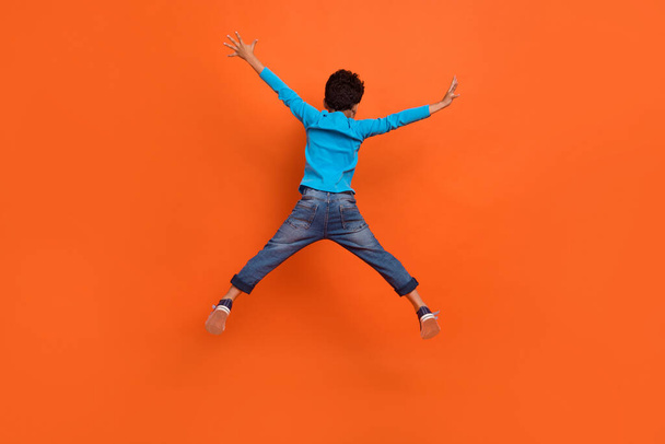 Full length photo of adorable small boy jumping back view spread legs arms dressed stylish blue clothes isolated on orange color background. - Foto, immagini