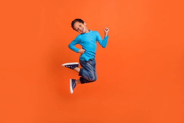 Full size photo of charming small pupil boy jump play raise fists celebrate dressed stylish blue look isolated on orange color background. - Fotó, kép