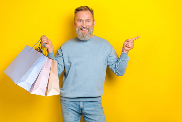 Portrait of handsome cheerful aged man hold packages indicate finger empty space isolated on yellow color background. - Foto, immagini
