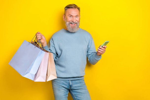 Portrait of positive aged man hold telephone boutique packages isolated on yellow color background. - Foto, imagen