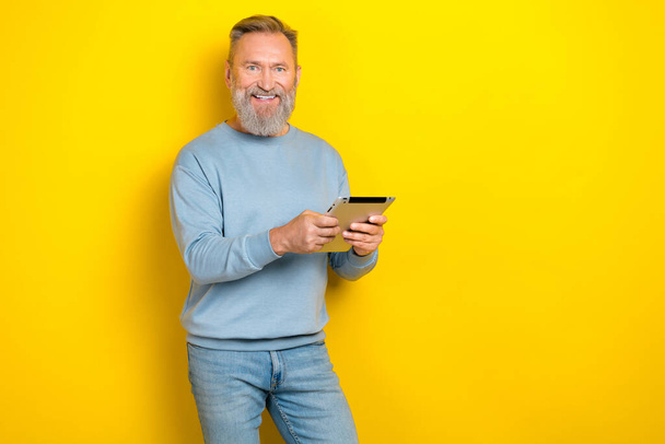 Foto de alegre hombre positivo pensionista vestido azul pulóver trabajando dispositivo moderno espacio vacío aislado color amarillo fondo. - Foto, imagen