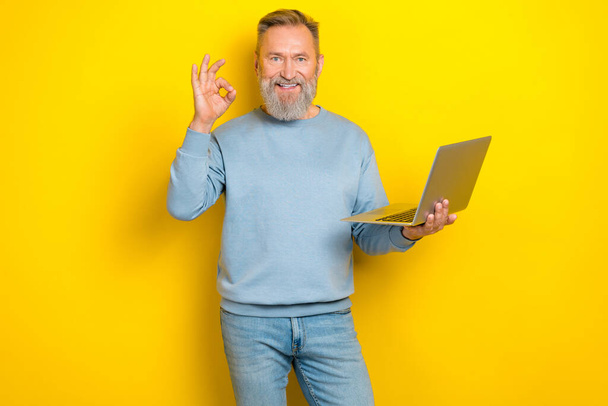 Photo of pretty cheerful man pensioner dressed blue pullover showing okey sign reading modern device isolated yellow color background. - Fotó, kép
